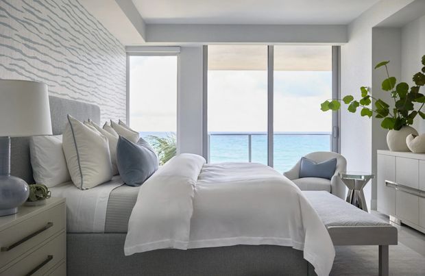 Bright, refined coastal bedroom with a grey upholstered bed dressed in layered white and pale blue bedding. The room features textured wave-pattern wallpaper, a contemporary bedside table and lamp, a low upholstered bench, streamlined white cabinetry, and full-height glazing opening to a balcony with ocean views. Natural greenery and sculptural decor add softness.