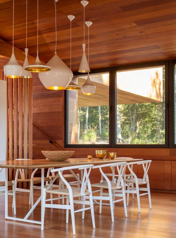 Warm, wood-lined dining room with a long timber dining table and white sculptural dining chairs. Multiple white pendant lights hang at varied heights above the table, while large dark-framed windows provide views into a wooded landscape. Built-in timber cabinetry and vertical timber detailing reinforce the natural, refined interior.
