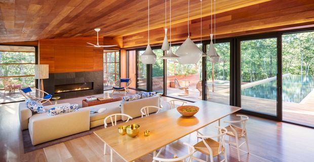 Open-plan contemporary living and dining area with extensive floor-to-ceiling glazing, warm timber wall and ceiling finishes, a linear fireplace, pale upholstered seating, and a large timber dining table beneath multiple sculptural pendant lights. A pool and landscaped greenery are visible outside through the glazing.