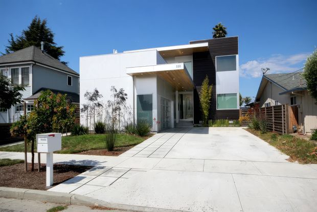 Street-facing view of a contemporary two-storey residential house with a crisp white facade, projecting flat-roofed entry canopy, dark vertical cladding panel, large rectangular windows, and a broad concrete driveway. The modern home is set among established neighbouring houses with a small landscaped front garden and clear blue sky.