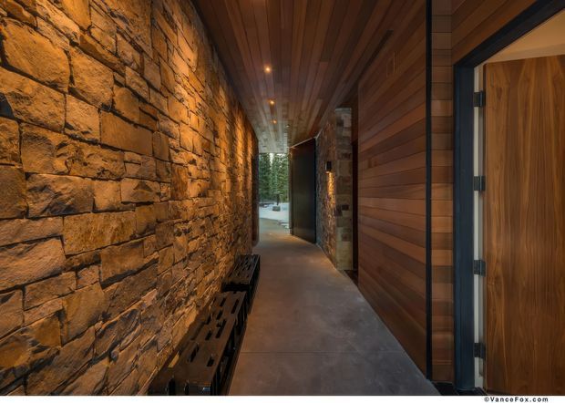 A narrow, atmospheric architectural corridor or entry passage with a rough stacked-stone feature wall on the left, dark timber wall cladding and a slatted timber ceiling, polished concrete flooring, integrated warm lighting, and a dark-framed timber door on the right. A bench runs along the stone wall, with a glazed opening at the end leading toward greenery.