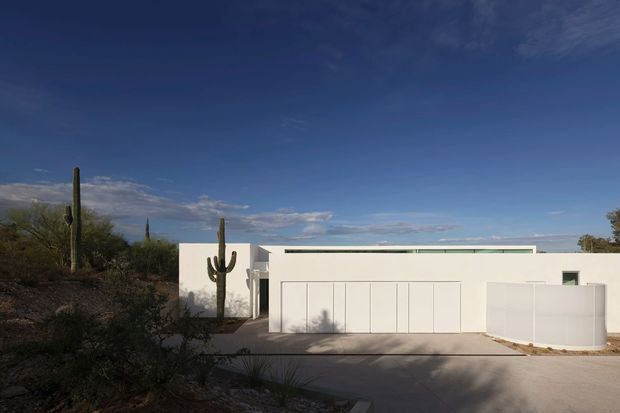 Low, minimalist modern residence with crisp white stucco-like walls, a concealed or very low roofline, and a broad garage or entry facade. The home is set in a dry desert landscape with tall saguaro cacti and sparse scrub, viewed from the driveway under a deep blue sky.