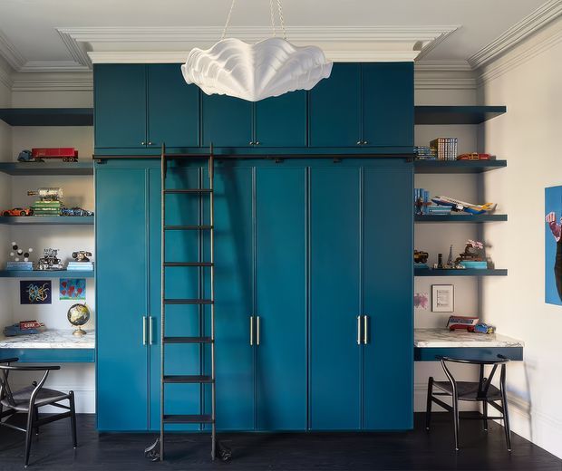 A highly styled children's or youth bedroom with a dramatic floor-to-ceiling teal built-in storage wall, integrated wardrobes, open shelving, desks, and a dark metal rolling ladder. The shelves display model vehicles, toys, books, and decorative objects. Two compact workstations sit on either side, beneath a sculptural white pendant light. The room features ornate white ceiling mouldings, dark timber flooring, and a playful yet refined design.