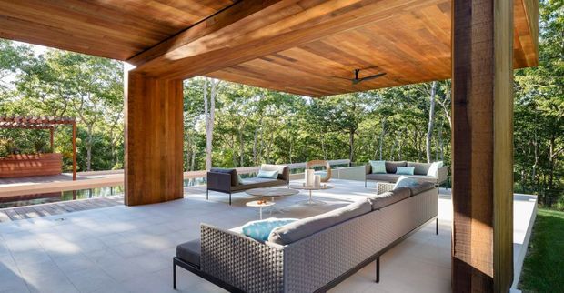 A spacious covered outdoor entertaining terrace surrounded by trees, with a large modular outdoor sectional, coffee tables, and additional lounge seating. The terrace features a substantial timber ceiling and posts, pale large-format flooring, and open sides overlooking the landscaped setting.