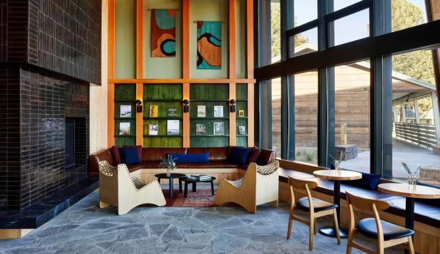 A double-height contemporary lounge or reception area with expansive black-framed windows, a dramatic dark tiled fireplace wall, built-in leather banquette seating, sculptural lounge chairs, small timber tables, and colourful abstract wall panels. The space has a warm, eclectic mid-century-inspired fit-out and a slate-like irregular stone floor, with an exterior deck and building visible through the glazing.