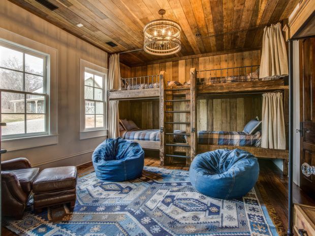 A warm, rustic bunk room with two levels of built-in wooden bunk beds, privacy curtains, a central ladder, blue beanbag chairs, leather seating, and a patterned area rug. The room features extensive reclaimed or weathered timber wall and ceiling panelling, large windows, and a decorative circular chandelier.
