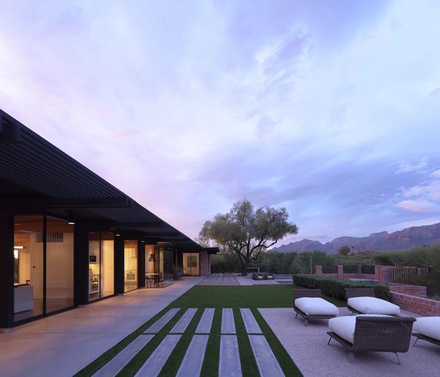 A low, modern single-storey residence opens onto a landscaped outdoor courtyard and lounge area. The broad dark roof overhang shelters full-height glazed rooms, while linear concrete or stone pavers are set into a manicured lawn leading toward gravel seating areas, clipped hedges, and a brick boundary wall. Mountainous terrain is visible beyond the property, under a soft blue and pink evening sky.