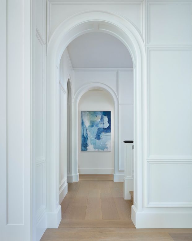 A bright, refined hallway with a sequence of ornate arched openings, white decorative wall panelling and moulded trims, and pale timber flooring. A blue abstract artwork provides a small colour accent at the end of the corridor, while a partial stair or landing edge is visible on the right.