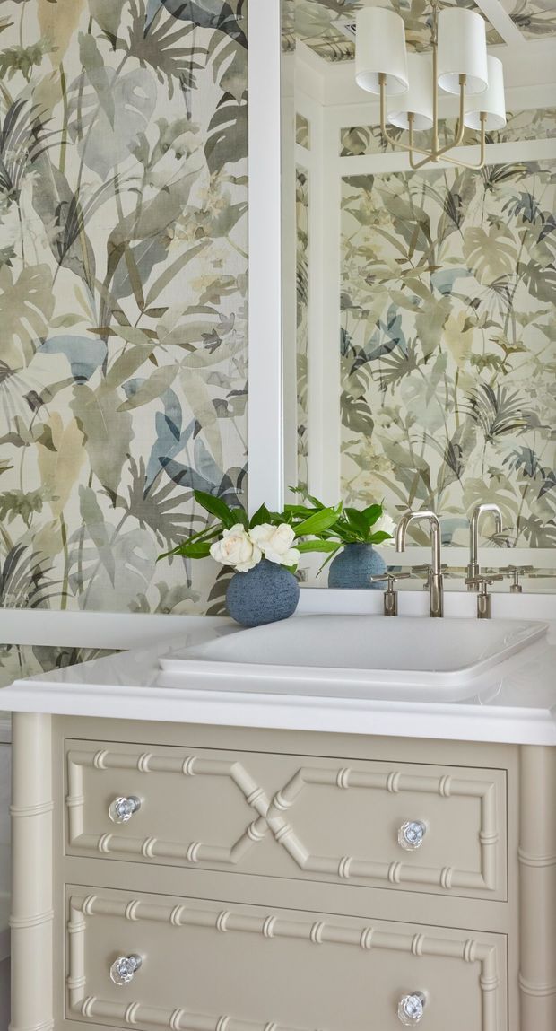 Close-up view of a refined bathroom vanity with a white vessel sink, polished chrome tapware, ornate cream cabinetry, and botanical-patterned wallpaper reflected in a large mirror. A sculptural multi-arm pendant light and small decorative plant arrangement add a soft, elegant character.