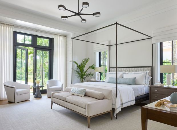 Bright, refined bedroom with a large black four-poster canopy bed, layered white bedding, upholstered bench, matching armchairs, dark timber bedside tables, tall black-framed windows and French doors, sheer curtains, and a contemporary sculptural ceiling light. The room has a calm neutral palette with tropical greenery visible through the glazing.