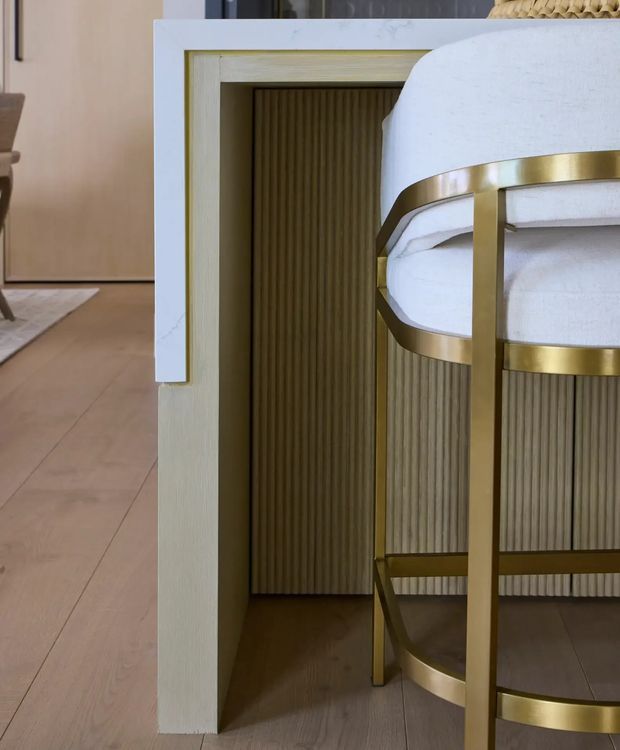 Close-up interior view of a refined dining or kitchen area featuring a light stone-clad peninsula or island with a recessed fluted timber-panelled base. A partially visible upholstered bar stool with brushed brass-gold framing sits at the counter. Warm timber flooring and neutral cabinetry create an elegant, softly layered interior.