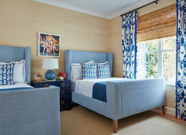 A bright twin bedroom with two upholstered blue beds, patterned blue-and-white bedding and curtains, a dark bedside chest with a lamp and decorative objects, woven-texture beige wallcovering, and a large window with a bamboo blind. The room has a coordinated coastal-inspired colour scheme and layered textiles.