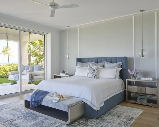 Bright, refined bedroom with a large upholstered blue-grey bed, white layered bedding, a matching upholstered bench, and a pale patterned area rug. The room features a paneled light-grey accent wall, timber flooring, slim pendant lights, a ceiling fan, contemporary bedside and storage furniture, and full-height glazing opening to a sunny outdoor setting.