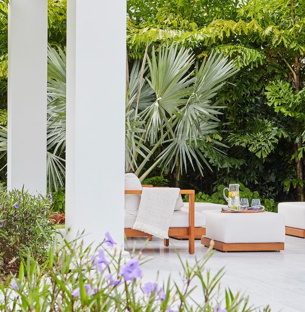 A bright tropical outdoor lounge or patio with white structural columns, pale tiled flooring, and contemporary white upholstered seating on timber bases. Dense palms and lush greenery surround the space, with a low table set for drinks and flowering plants in the foreground.