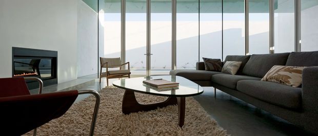 A contemporary living room with a large dark sectional sofa, low oval glass coffee table, shag rug, sculptural lounge chairs, and a built-in linear fireplace. Floor-to-ceiling glazing opens to expansive mountain views, creating a bright, refined interior with strong indoor-outdoor visual connection.