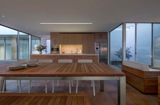 Open-plan contemporary kitchen and dining area with extensive floor-to-ceiling glazing, warm timber cabinetry, a large slatted timber dining table and bench, pale upholstered dining chairs, and a timber-framed built-in bench seat. The room has a clean, minimal composition, polished concrete or stone flooring, and abundant natural light.