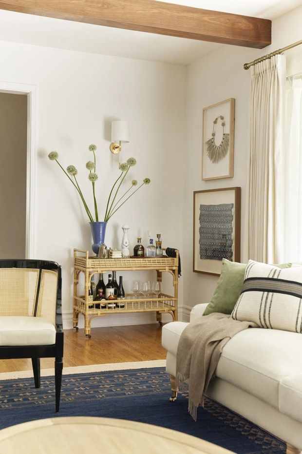 A bright, refined living room with warm white walls, polished timber flooring, and a prominent exposed timber ceiling beam. The room includes a white sofa with green and striped cushions, a black-framed cane chair, a woven rattan bar cart stocked with bottles and glassware, framed textile and decorative artwork, brass wall lighting, full-height cream curtains, and a navy patterned area rug. The palette is warm and inviting with traditional and natural woven accents.
