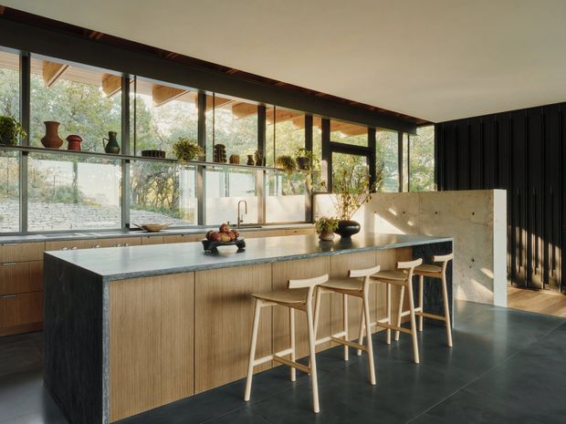 A spacious contemporary kitchen with a large central island, textured wood cabinetry, a dark stone benchtop, and four timber bar stools. Full-height glazing runs along the rear wall, bringing in abundant natural light and views of greenery. A concrete-toned partition and dark vertical wall cladding add contrast, while decorative shelving and potted plants provide warmth.