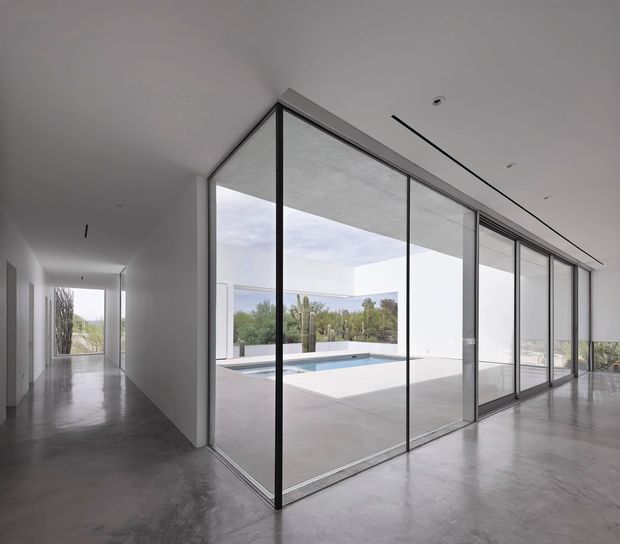 A minimalist contemporary interior with a polished grey concrete floor, white walls and ceiling, and a dramatic full-height glazed corner overlooking a courtyard swimming pool. Slim dark-framed sliding glass doors create a strong indoor–outdoor connection, while a hallway extends along the left side. The space is bright, open, and highly restrained, with recessed ceiling fixtures and a linear ceiling slot detail.