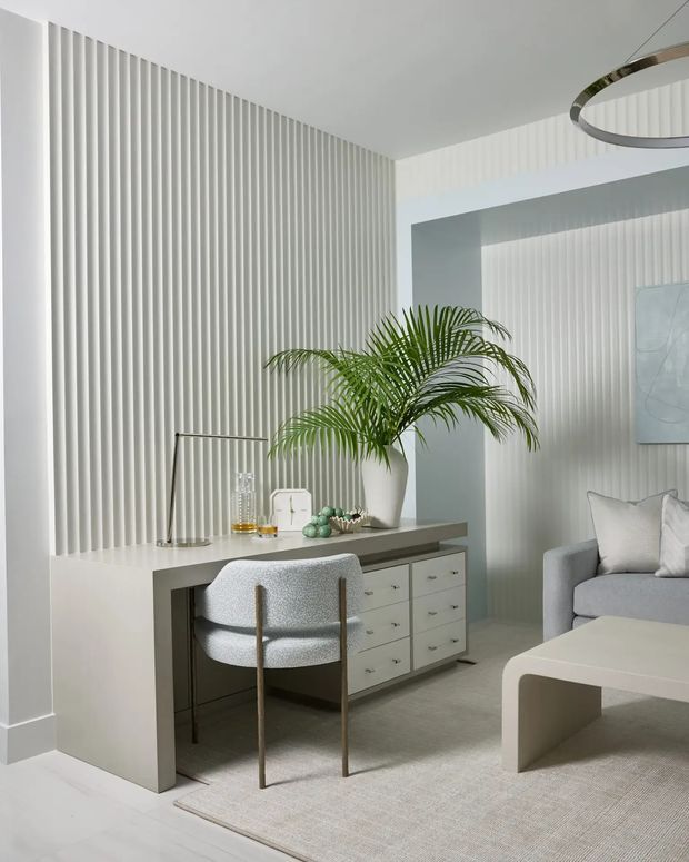 A bright, refined contemporary interior combining a compact home workspace with a living area. A slim console desk with drawers sits against a full-height white fluted wall, paired with a rounded upholstered desk chair and simple decorative objects. A potted palm, pale sofa, curved off-white coffee table and neutral rug create a soft, minimalist atmosphere. A sculptural circular pendant is partially visible overhead.