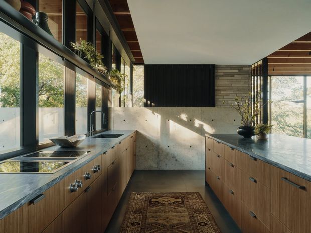 A warm, contemporary galley kitchen with extensive floor-to-ceiling glazing, timber cabinetry, dark stone benchtops, and a central walkway. A black vertical-panel feature wall and stacked-stone wall add texture, while integrated shelving, indoor plants, and a patterned runner create a refined natural atmosphere.