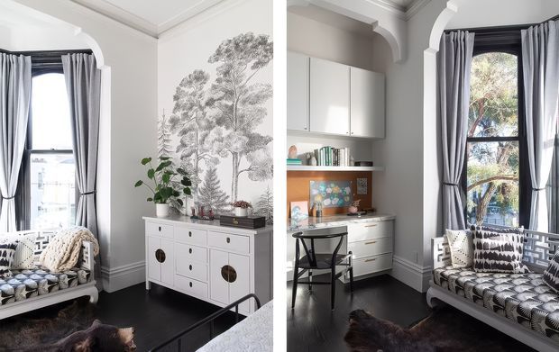 A bright, refined interior combining a sitting area and compact home office. Tall arched windows are dressed with grey curtains, while white built-in cabinetry, a small desk, dark timber flooring, decorative wall moulding, and a patterned upholstered settee create a classic yet contemporary character. A large monochrome botanical wall mural and leafy plant add visual interest.