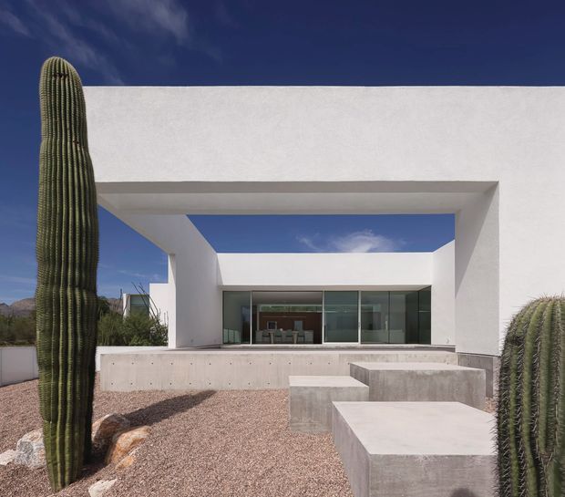 A minimalist modern single-storey residence with a stark white stucco facade, deep rectangular portal opening, extensive glass doors, and a raised concrete terrace. The desert landscape features gravel landscaping and tall columnar cacti, with rugged mountains visible in the distance beneath a deep blue sky. Raw concrete steps and block seating lead toward the sheltered outdoor living area.