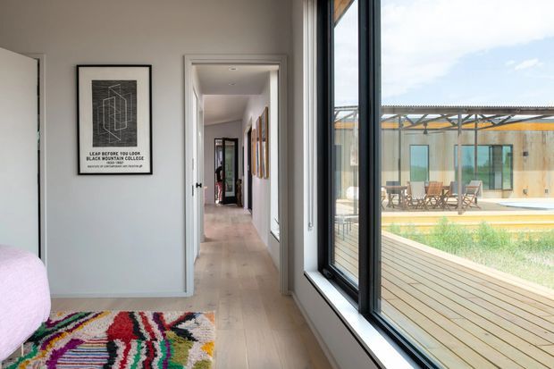 Bright contemporary interior view from a bedroom or adjoining hallway, looking along a white-painted corridor with pale timber flooring and framed artwork. A large black-framed window wall opens the space visually to a timber deck, swimming pool and a low modern building outside. The foreground includes part of a bed and a colourful patterned rug, while the hallway extends toward additional rooms and a mirror.