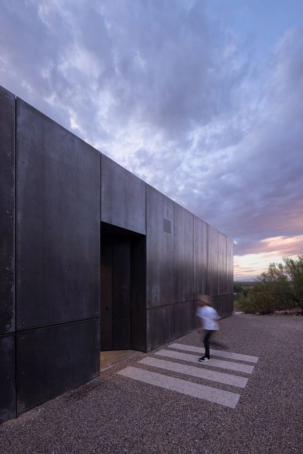 Exterior architectural photograph of a stark, rectilinear building clad in dark charcoal metal panels. A recessed entry door interrupts the long monolithic facade, approached by broad rectangular concrete stepping slabs set into a gravel landscape. The scene is photographed at dusk, with dramatic purple-blue clouds, distant desert-like terrain and sparse shrubs visible beyond the building. A blurred person provides scale.