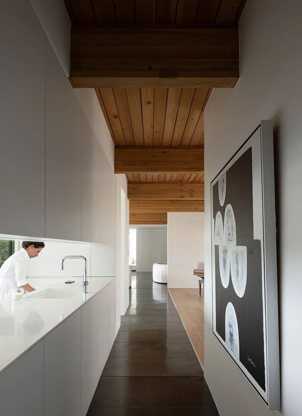 A refined open-plan residential interior viewed along a narrow passage beside a white minimalist kitchen. The kitchen has tall white cabinetry, a white island with an integrated sink and chrome tap, and a person preparing food. The corridor leads toward a bright living area, with a timber-lined ceiling and substantial exposed timber beams overhead. Dark timber flooring continues through the space, while a large monochrome artwork is mounted on the right wall. The overall design combines clean contemporary joinery with warm natural timber detailing.