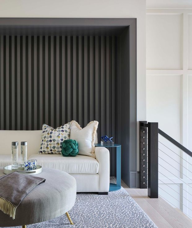 Refined contemporary living room with a white upholstered sofa positioned in front of a dramatic black vertically ribbed feature wall. A rounded light-grey ottoman, patterned cushions, small side table, decorative objects and a pale patterned rug create a soft contrast. The room opens to a stairwell with white balustrade panels and dark timber or painted posts, while built-in wall mouldings add architectural detail.