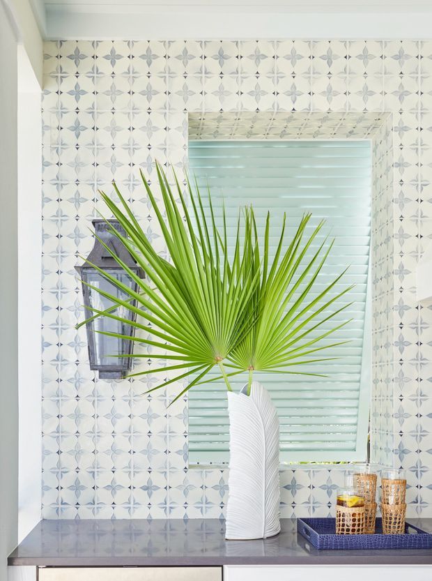 Bright interior vignette featuring a patterned blue-and-white tiled wall, a large louvred window with pale horizontal shutters, and a dark grey stone benchtop. A sculptural white vase with fan palm leaves and a blue tray with woven drinking glasses provide decorative accents. The scene has a fresh, tropical-coastal feel with abundant natural light.