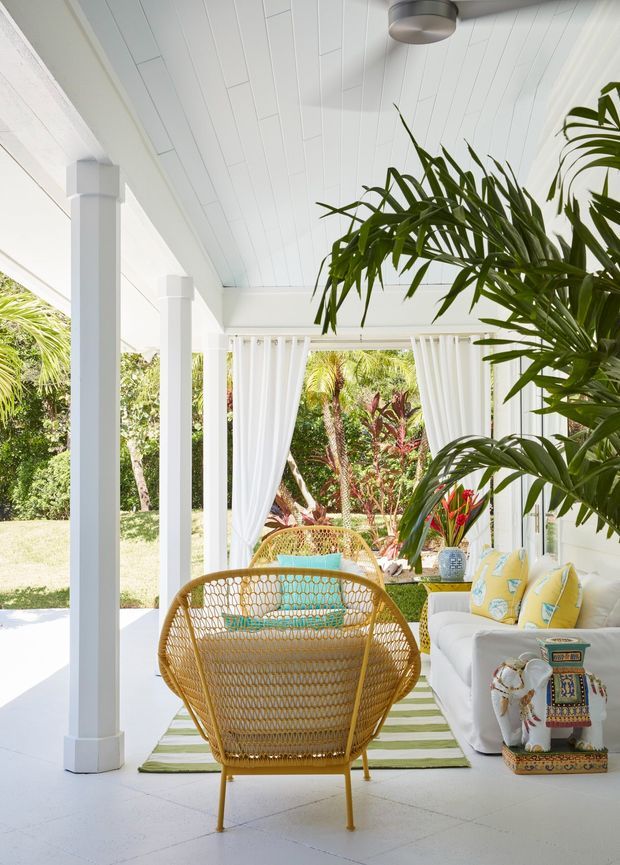 Bright tropical enclosed veranda or sunroom with white-painted columns, walls, ceiling boards and tiled floor. Sheer white curtains frame an open garden view, while rattan lounge chairs, a white sofa with patterned cushions, indoor palms and colourful decorative accents create a relaxed resort-like setting. A ceiling fan and extensive greenery reinforce the airy tropical character.