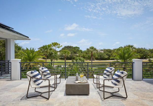 A bright elevated outdoor terrace overlooking landscaped greenery and a waterway or golf-course-like open setting. The terrace has pale stone or concrete paving, a dark horizontal metal balustrade with white rendered posts, and four woven outdoor lounge chairs arranged around a square concrete coffee table. A partial modern roofline is visible at left under a clear blue sky.