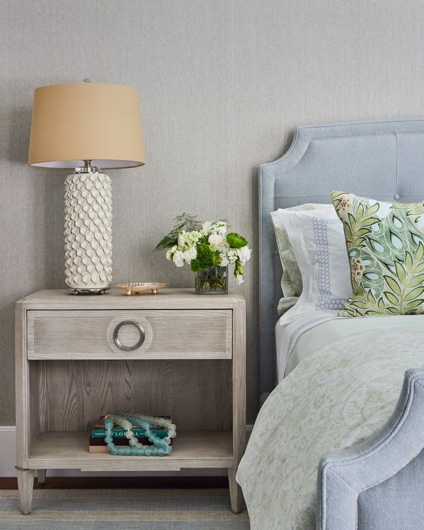 Close-up of a softly styled bedroom corner featuring a light blue upholstered bed with a curved, buttoned headboard and footboard, a greyed timber bedside table with circular drawer hardware, a textured ceramic table lamp, layered white and patterned bedding, and a small floral arrangement. The room has a calm, refined, traditional-contemporary feel with a neutral grey textured wall and carpeted floor.