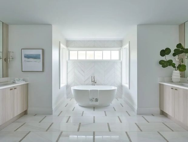 Bright, symmetrical contemporary bathroom with a freestanding white bathtub positioned beneath a bank of clerestory windows. The room features pale marble-look tiled walls and floor with linear contrasting insets, light timber floating vanities on both sides, large mirrors, and brushed metal tapware. A potted plant and framed artwork add restrained decorative accents.