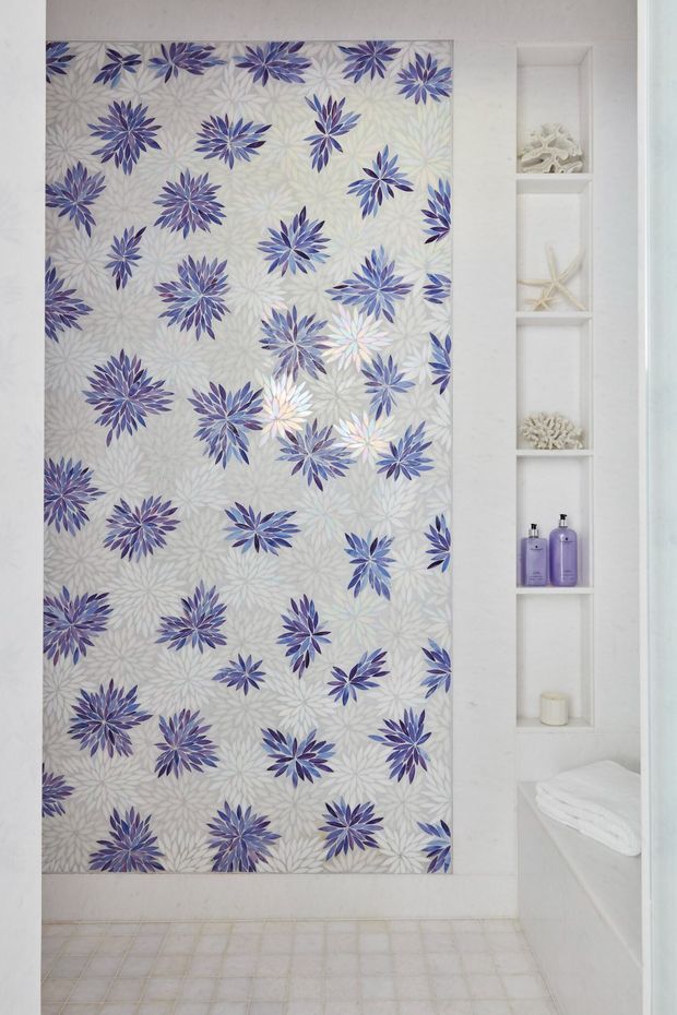 Bright white bathroom featuring a full-height floral-patterned shower curtain or screen in soft grey, lavender, and blue. A narrow recessed shelving niche displays shells, coral-like ornaments, toiletries, and a candle. The room includes pale tiled flooring, white walls and built-in joinery, with folded white towels visible at right.