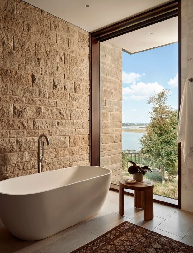 A luxurious bathroom with a freestanding white bathtub positioned beside a full-height glazed opening. The room features a textured tan masonry feature wall, pale tiled flooring, dark timber window and door framing, and a brushed metal floor-mounted bath tap. A small timber side table with a plant and a patterned rug add warmth. Through the glazing is a bright waterfront landscape with water and sky.