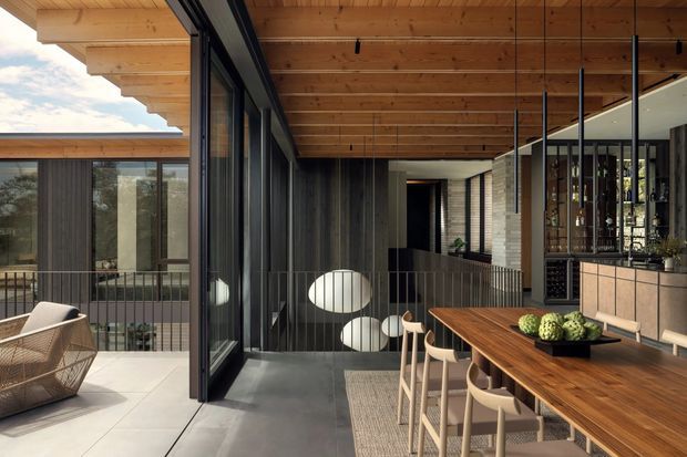 A sophisticated covered outdoor dining terrace connected to a contemporary home, featuring a large timber-lined soffit, dark-framed full-height glazing, concrete flooring, a long timber dining table with pale chairs, and a built-in bar or kitchen zone. The space overlooks an internal courtyard or lower-level area and uses a restrained palette of timber, charcoal, concrete, and light stone.