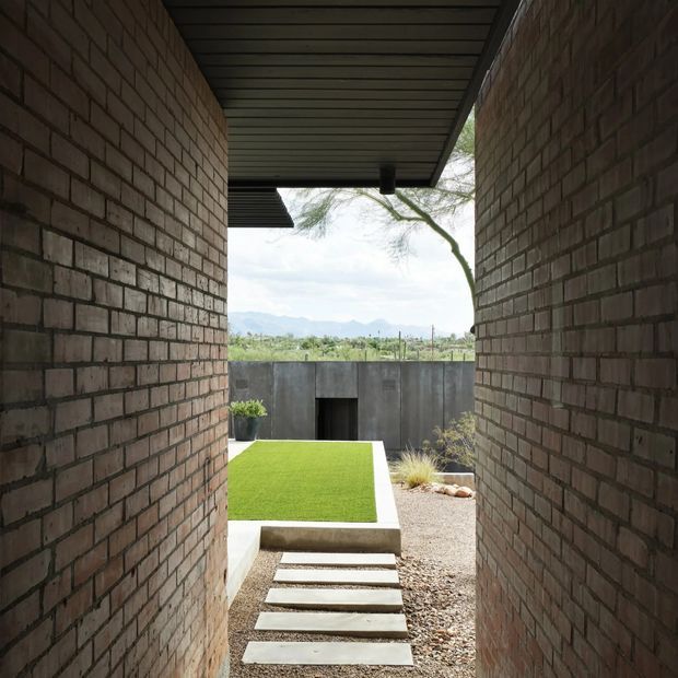A narrow covered passageway framed by exposed brick walls and a dark slatted ceiling opens onto a landscaped courtyard. Large rectangular concrete stepping stones cross a gravel surface toward a raised bright-green lawn, with a concrete boundary wall and distant mountain landscape visible beyond.