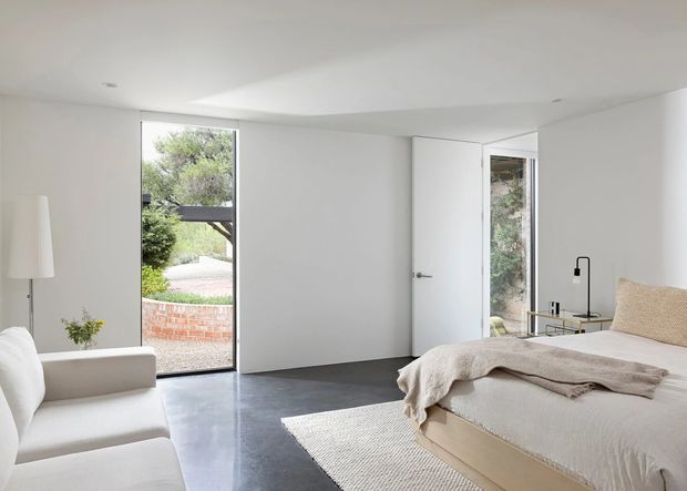Bright minimalist bedroom with white walls and ceiling, a low-profile upholstered bed, pale neutral bedding, a white sofa, and a woven rug. Floor-to-ceiling glazing and an open glass door provide views to a landscaped exterior with a brick garden wall. Dark polished-looking flooring creates contrast with the light interior, while simple recessed lighting and a slender black lamp add understated detail.