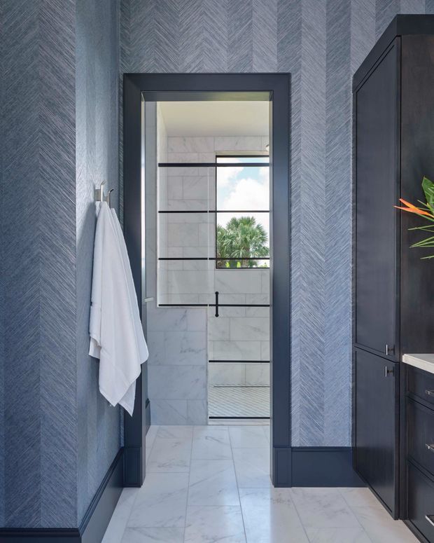 A refined bathroom interior viewed from a darker blue-grey vanity area, looking through a framed opening into a marble-tiled shower enclosure. The space features herringbone-patterned blue-grey wallcovering, dark navy or charcoal cabinetry and trim, white marble-look floor and wall tiles, black-framed shower glazing, and a narrow outdoor view through the shower window. A white towel hangs from a wall hook, while a partial vanity and botanical accent appear at right.