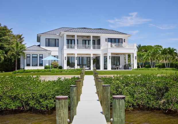 A large two-storey white coastal-style residence viewed from a private dock or boardwalk. The home has a symmetrical facade, broad covered balconies with white railings, tall glazed doors and windows, white columns, and a low-pitched grey metal hipped roof. Manicured hedges and palms surround the property, with water in the foreground and a walkway leading directly to the house.