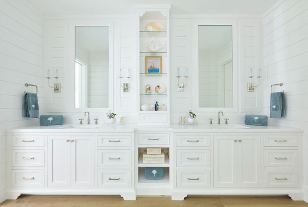 Bright, symmetrical double-vanity bathroom with extensive white painted cabinetry, two rectangular mirrors, a central open display niche, paneled walls, twin basins, and polished metal fixtures. Soft blue towels and small decorative accessories provide subtle contrast, while a light timber floor adds warmth.