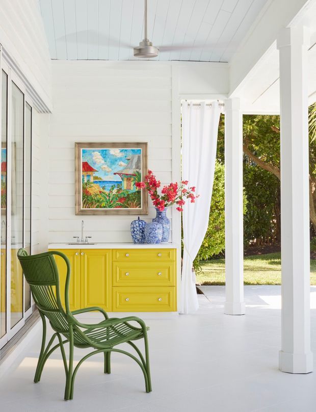 Bright covered outdoor veranda or sunroom with white-painted horizontal wall panelling, a white boarded ceiling, white structural columns, and large glazed openings. A vivid yellow built-in sideboard with sink sits beneath a framed tropical painting, accompanied by blue-and-white ceramic vases and a green wicker lounge chair. White curtains open to a leafy garden, creating an airy coastal, tropical atmosphere.