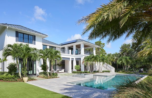 Bright exterior view of a large two-storey white residence with horizontal cladding, dark pitched metal roofs, broad covered verandas, columns, and extensive glazing. A paved terrace and swimming pool occupy the foreground, with tropical palms and landscaped garden surrounding the house.