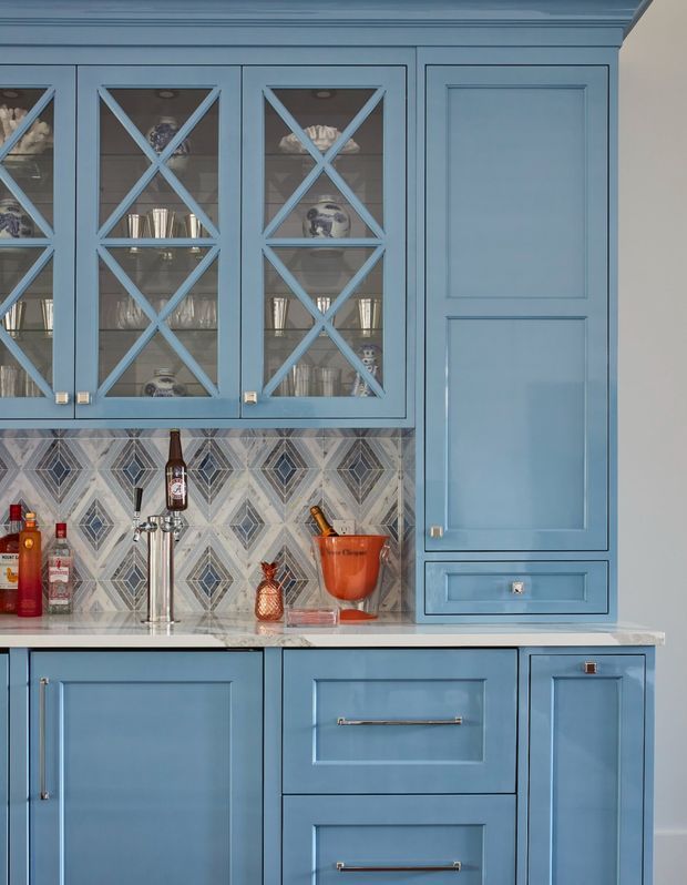Close-up view of a blue built-in home bar or beverage station with shaker-style cabinetry, glass-front upper cupboards featuring decorative X muntins, a patterned geometric tile splashback, pale stone benchtop, and a polished metal tap. Bottles, glassware, and bar accessories are visible.