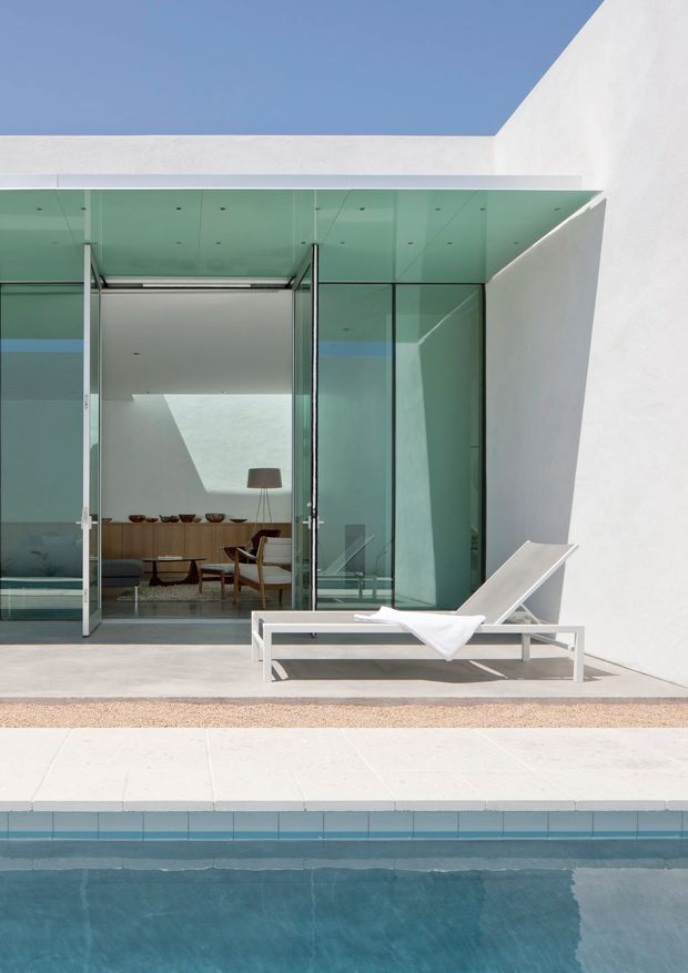 A minimalist modern poolside terrace with a white rendered geometric facade, expansive floor-to-ceiling glass sliding doors, and a pale tiled pool surround. A white sun lounger sits beside the swimming pool, while a refined dining and living area is visible through the glazing. The composition features strong clean lines, abundant natural light, and a restrained white, pale blue, and turquoise palette.