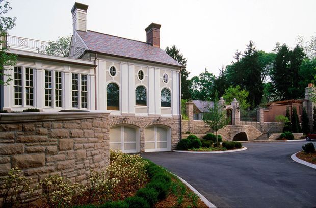 Exterior view of a formal multi-level stone-and-plaster residence or estate. The building features a steep slate-clad roof, prominent chimneys, tall white-trimmed arched windows, classical detailing, and a projecting glazed wing. Stone retaining walls, landscaped planting, gates, and a paved driveway frame the property.