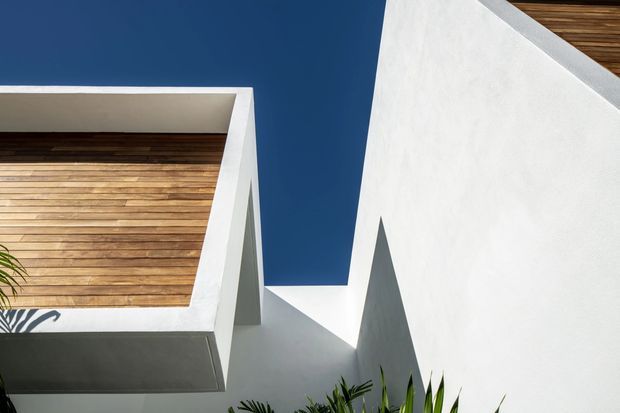 Abstract close-up architectural exterior showing crisp white rendered walls and projecting geometric forms around a recessed balcony or terrace. Warm horizontal timber lining is visible beneath the overhangs, with strong sunlight creating angular shadows against a deep blue sky and partial tropical planting at the bottom edge.